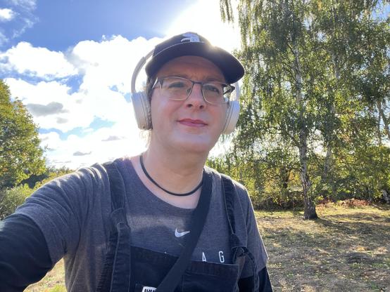 A selfie of Ellie, who’s sitting on a bench outside in front of some grass and trees on a sunny day. 

She’s looking at the camera and is wearing a Chicago White Sox cap, white headphones, glasses, a black necklace, a grey Chicago White Sox t-shirt (am I dangerously close to full kit wanker territory here?) with a black long sleeve top underneath, and black dungarees.