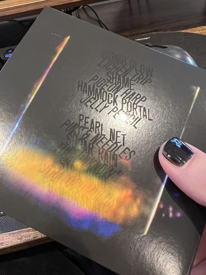 A photo of the back cover of the Bibio & Dorian Concept collaboration album Answers. The image is a photo of what looks like a black box on a black background with only the side edges visible and a rainbow coloured cloud effect at the bottom. The track listing is printed with spot laminate so it’s only visible by tilting the CD and catching the light at the right angle.