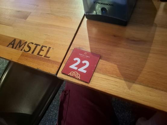 Mesa de bar dw madeira na qual está escrito a marca da cerveja (Amstel) em preto e o número 22,  escrito em branco num adesivo quadrado vermelho