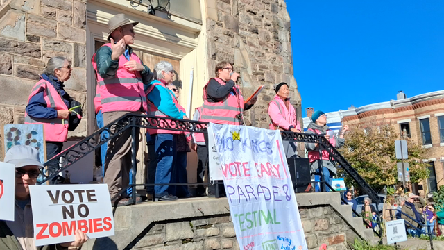 People assembled around and on some church steps

Signs:

47 xed out

Vote no zombies

No kings vote early parade & festival