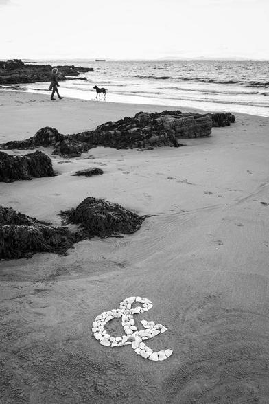 A black and white photo. A white ampersand made of shells is on a sandy beach. Some dark rocks behind the ampersand point off towards the sea, where a labrador and their human are silhouetted against the water's edge.