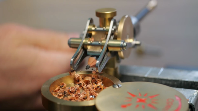 A short sequence shows the pencil sharpener in action. The shot is tightly focused on the small machine, which is made from polished brass and steel, and driven by an out-of-focus human hand that turns a hand crank.The pencil is held in the middle of the machine, and a blade, which is driven by the hand crank along two guide slots at each side of the pencil, shaves off small pieces of wood and graphite at an angle, like when sharpening a pencil with a pocket knife. The pencil is also slowly rotated along its axis by the mechanism, so that all sides are being cut. Under the pencil sits a round brass tray for collecting the chips.