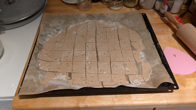 The dough has been flattened very thin with a rolling pin on a sheet of parchment and a baking sheet, and divided into small rectangles. The brown caraway seeds are poking through.