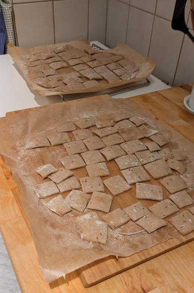 two sheets of parchment paper covered with small, brown, rectangular, slightly floury bread crackers cooling off on the kitchen counter, dark brown caraway seeds poking through on the top