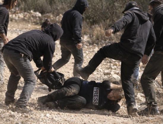 A group of people in dark clothing are gathered around a person lying on rocky, dirt ground. The person on the ground is wearing a dark vest with the word "PRESS" printed in white. One person in the foreground leans over the person on the ground near black equipment. Another person has a leg raised toward the person on the ground with a clenched fist. The background consists of a dry, hilly landscape with sparse, leafless vegetation.