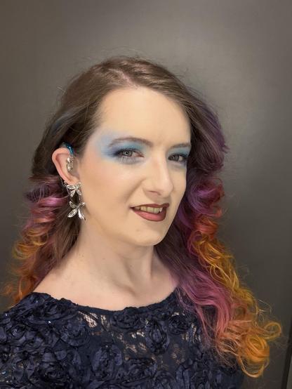 Hazel looks at the camera with blue eyeshadow, flower earrings, red lipstick, and curly sunset coloured hair. She’s wearing a blue dress