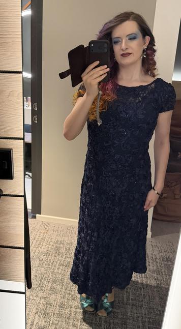 Hazel looks at the camera with blue eyeshadow, flower earrings, red lipstick, and curly sunset coloured hair. She’s wearing a blue dress and sparkly blue heels