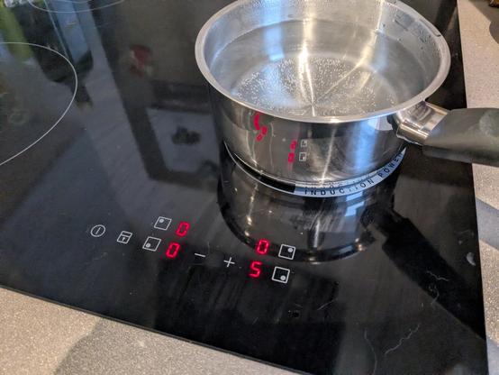 induction cooktop with capacitive controls and four seven-segment LED displays reading 0, 0, 0, and 5. a saucepan of water coming to boil on the active hotplate