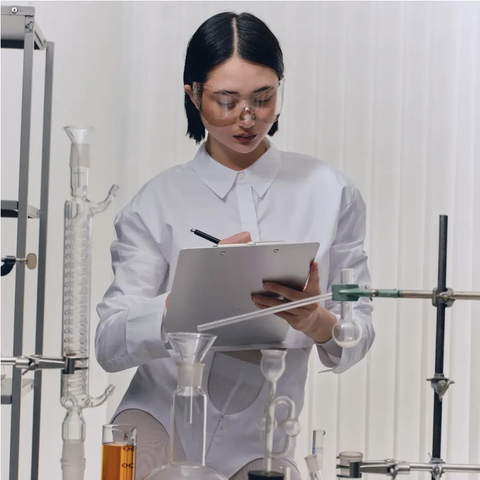 AI generated slop of a woman making notes on a pad, in a lab. There are clamp stands with some glass devices used typically in a biochemical labs. The woman wears some protective glasses, who are rather a cyberpunkish fashion statement, that being fit for the purpose, as they are cut off in a way that makes it possible to get splashes in the eyes. She wears white pantyhoses, and a white body. The body has a cutoff around the navel, showing the waist line of the pantyhoses underneath. There's a zipper on the bottom part of the body, below the cutoff. The top of the cutoff is not visible, as it's hidden beyond the notepad. It'd hard to determine it for sure on this slop, but it looks like there is another garment worn over the body. It's a white office shirt, with wide cuffs, and a collar, but it's rather short, ending on the waist line, leaving the cutoff in the body exposed. It looks like a feverish dream of sexy scientist. My eyes are bleeding. I want to lobotomise myself with an icepick to forget this cursed view.