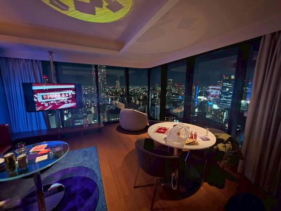 a hotel room lit up with mood lighting and a reflective mirror table and a ceramic table with drinks and shopping on it and a tv on and chairs and a panoramic night view of Osaka