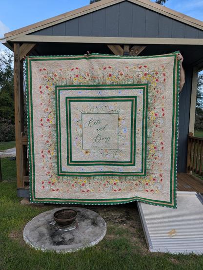 A large, bed-sized quilt hanging in front of a small, "portable" building. The quilt is square, with green, ivory, and gold borders, along with border prints of wildflowers in red and yellow. The center medallion, also a square, has text "Kate and Doug October 18, 2026", which is surrounded by a square green frame and more wildflowers. The binding is a striped binding of the green, ivory, and gold stripes.