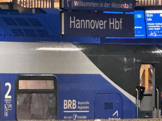 An einem Bahnhof steht ein Zug an einem Gleis. Auf der Bahnhofstafel steht „Hannover Hbf“ und „Willkommen in der Messestadt“. Am Zug darunter steht „BRB Bayerische Regiobahn“.
