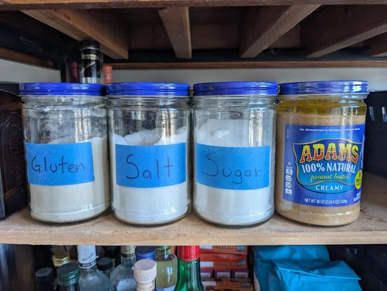Row of large jars. On the far right is an unopened jar of Adams peanut butter. To the left of it are the same jar but filled with dry goods, in right to left order: sugar, salt, gluten.