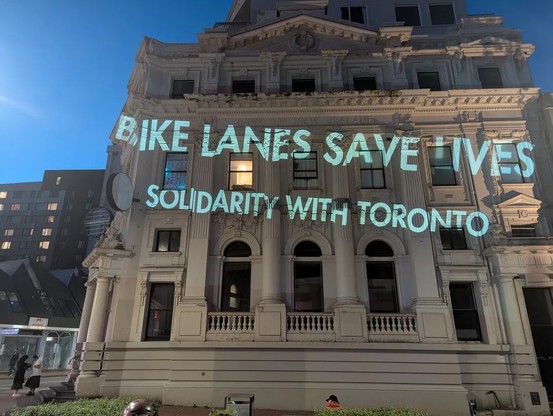 A public building at night with a light projection on the facade which reads:

"BIKE LANES SAVE LIVES - SOLIDARITY WITH TORONTO"

The photo is by Alex Dyer and the text is by Tom Flood