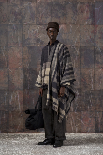 A model wearing one of the designs. It's a loose fitting top garment which is draped and has an oversized pattern that is not quite tartan in black, cream and grey. Loose black trousers underneath. It's paired with a beanie, black dress shoes and a large handbag.