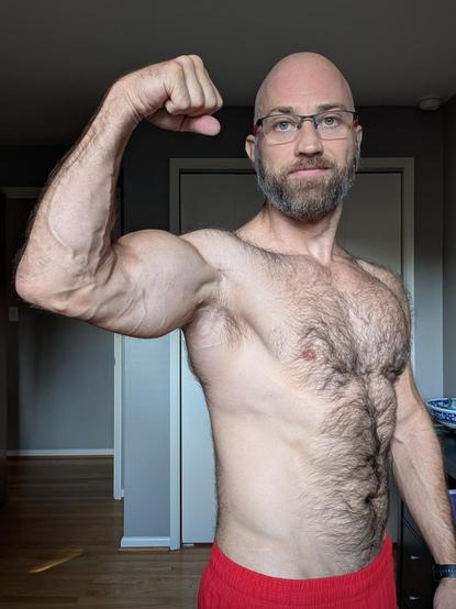 Shirtless, furry-bodied white guy stands in a home office flexing his right raised arm.