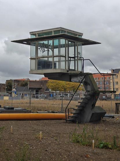 A photo showing a small elevated glass structure with a flat roof on a concrete base, a metal staircase leading to the entrance. The structure stands in an empty area inside a city. One can see a large yellow pipe lying on the bare dirt.