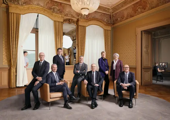 Formal group portrait of eight Swiss officials in suits and formal clothes, posing around tan armchairs in an ornate salon with gold-patterned walls, elaborate ceiling mouldings, huge curtained windows and a crystal chandelier. The production of the portrait is visible around its edges: in the large mirror behind the group, two crew members manipulate a bright octagonal light box, including one standing atop a stepladder. Another crew member is partly hidden behind a curtain to the left of the image. Through an open doorway on the right, a seated operator faces a computer monitor displaying the same group portrait. The polished official image deliberately contains much of the apparatus and labour involved in making it.
