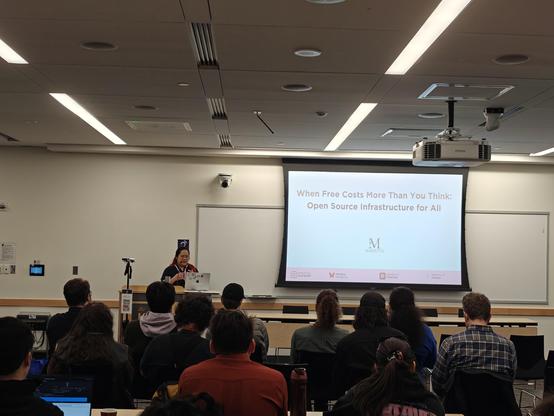 A person at the front of a large classroom full of people next to a screen that reads, "When Free Costs More Than You Think: Open Source Infrastructure for All"
