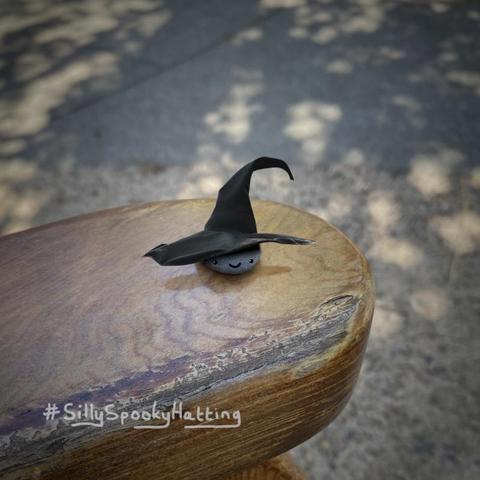 Original alt txt: A round dark-gray pebble on the end of an outdoor bench’s armrest, with tree-dappled shade on a sidewalk beyond
My addition: The image has been reframed into a square with a slight vignette on it. The pebble now has a sweet smiling face drawn on, and a very very wide brimmed witches hat.