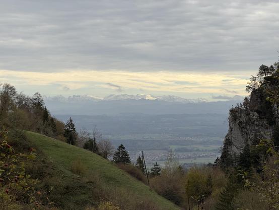 Bewölkter Himmel, darunter eine Alpenkette und die Sicht ins Tal, links und rechts Hügel und Felsen mit Bäume und Wiese
A cloudy sky, with a range of Alps below and a view of the valley; to the left and right, hills and rocks with trees and meadows