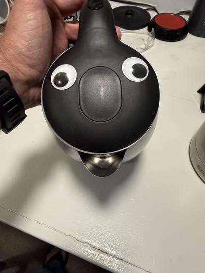 A black electric kettle as seen from above, the view looking down at the lid. Visible are two large-ish googly eyes set on either side and below the handle in the center of the lid is an oval section which resembles a nose. At the bottom is the stainless steel spout, which seems to form an open mouth. The overall effect is that of a cartoonish, anthropomorphized kettle (I’m a little teapot?).