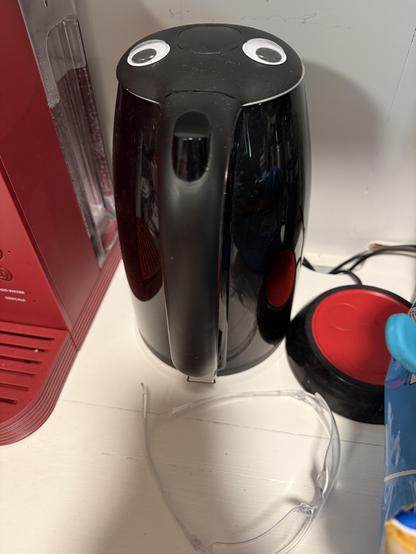 A black electric kettle on its base. On the lid are affixed two googly eyes. To the left of the kettle is a red Keurig coffee maker, and to the right is a mug warmer with a red warming disk. There is a pair of clear plastic safety glasses resting in front of the kettle.