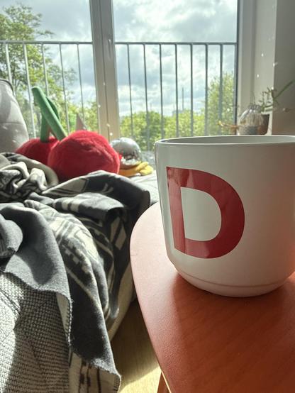 A coffee Cup with a large red letter „D“ on it in front of a window 