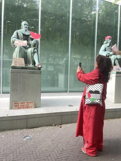 Helemaal in het rood gekleed neemt een vriendin een foto van een van de beelden bij het gebouw van de Hoge Raad. Het bronzen beeld is voorzien van bordjes zoals “Free Palestine”