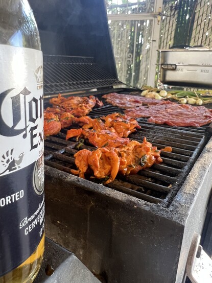 Photo of a grill with a beer in the foreground and chicken, carne asada, and onions cooking. 
