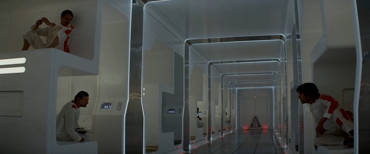 Screenshot of TV Show Andor in the prisoner hallway with inmates watching the door from their cells. All white futuristic small individual living space and an electrified floor. 