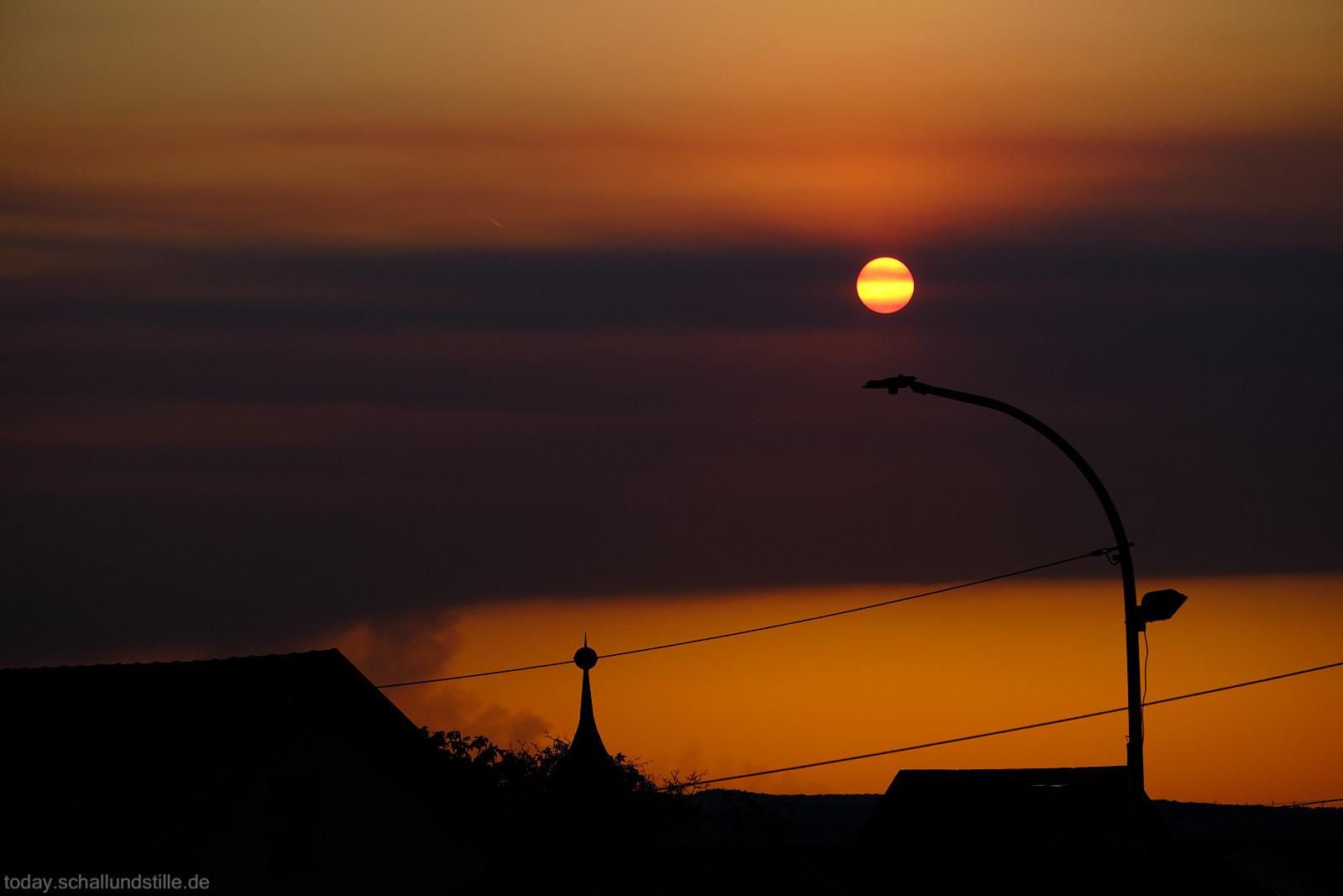Im Vordergrund sind die Silhouetten von Gebäuden und einer Straßenlaterne zu erkennen. Der Himmel ist von einem intensiven orangefarbenen Licht durchzogen, das von der tiefstehenden Sonne ausgeht. Über dem Horizont ziehen dunkle Rauchschwaden, die als Wolken erscheinen, jedoch tatsächlich vom Waldbrand in der Eifel stammen. Links im Bild ist deutlich die Rauchfahne sichtbar, die sich vom Horizont nach oben erstreckt.