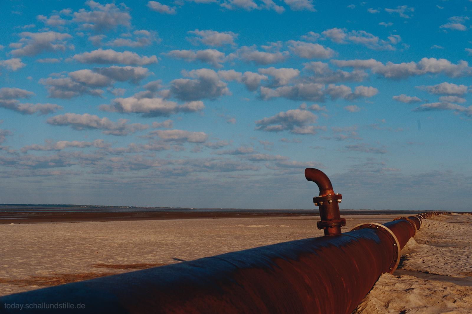 A large metal pipe extends across a sandy landscape toward the horizon. The pipe has a curved outlet near the foreground. The sky is bright blue with scattered white clouds. The scene is open and flat, with no vegetation visible.