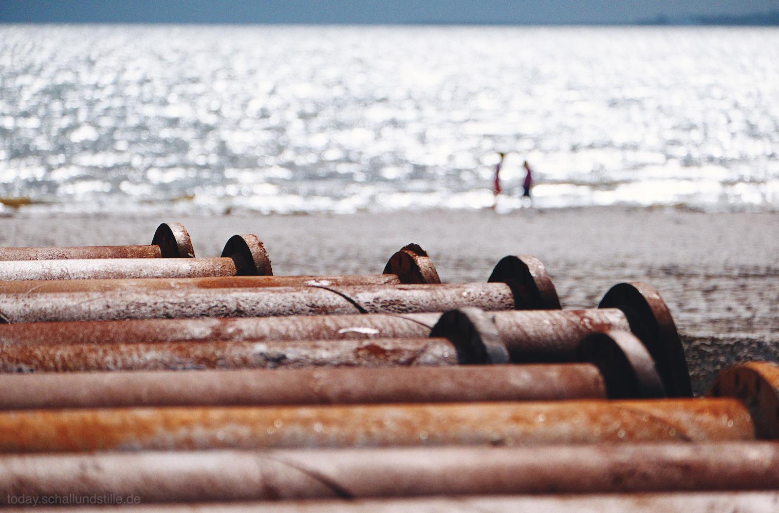 Rusty metal pipes lie in a row on the foreground, their surfaces weathered and worn. In the background, two blurry figures stand near the shoreline, appearing small against the vast expanse of water. The setting is a beach with the ocean stretching into the horizon. The light suggests a sunny day, with reflections shimmering on the water's surface. The image is framed to focus on the pipes in the foreground, with the background slightly out of focus, creating a sense of depth and distance.