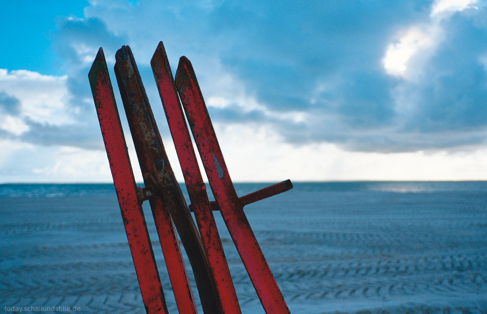 Four weathered metal posts, painted red and showing signs of rust and wear, stand upright in the foreground. The posts are positioned on a wide beach with tire tracks in the sand, leading toward a calm sea under a blue sky filled with large white and grey clouds. The scene is captured in bright daylight with a high-contrast color palette of red, blue, and white. The photograph is a close-up shot with a shallow depth of field, making the metal posts sharp while the distant ocean and horizon are slightly blurred.