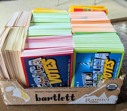 A cardboard bartlett pear box is full of postcards that are ready to mail. The cards are different bright colors and will be delivered all over the US.