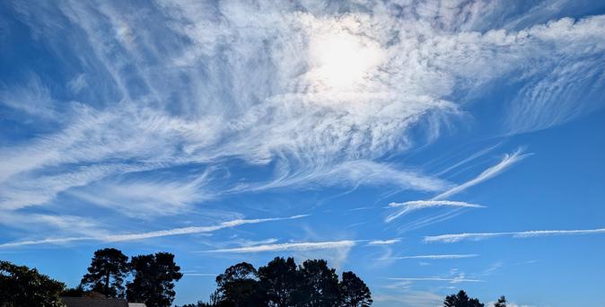 An intensely blue sky with wisps of clouds going all directions. The sun is behind the clouds. Dark tree tops are barely showing at the bottom.