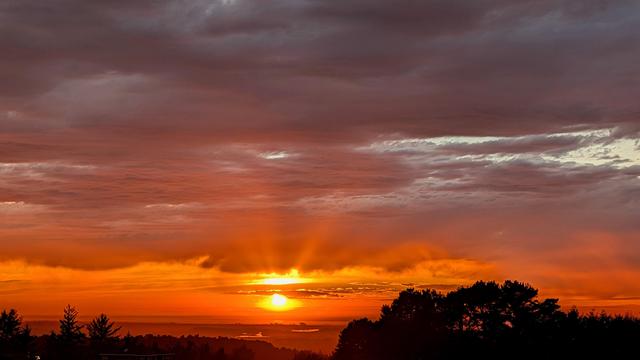 The sun is sending up feelers as it shines between the clouds. The darker clouds above look like rain. Dark tree tops at the bottom.