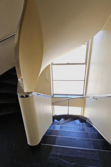 View down a modernist staircase towards a window. The walls are cream, the handrail is chrome, and the carpet is blue.