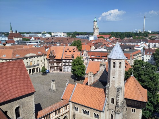 View from a tower onto a square with half-timbered buildings and a bronze sculpture of a lion in the middle. More modern buildings can be seen in the background and the chimney of a power station is far away. The weather is sunny and the sky is very blue.