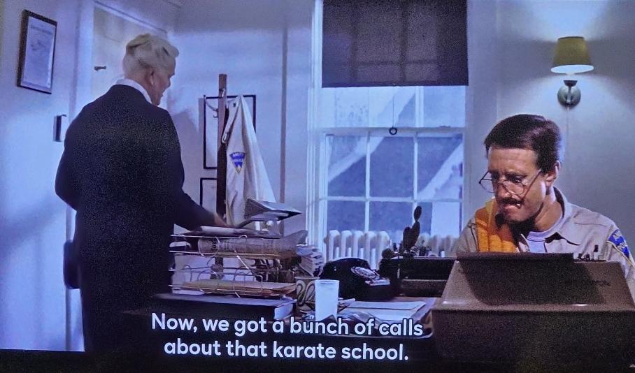A grey haired woman in a business suit is going through the inbox on the desk by which Roy Scheider in a sheriff's shirt is typing at a typewriter. She tells him, "Now, we got a bunch of calls about that karate school."