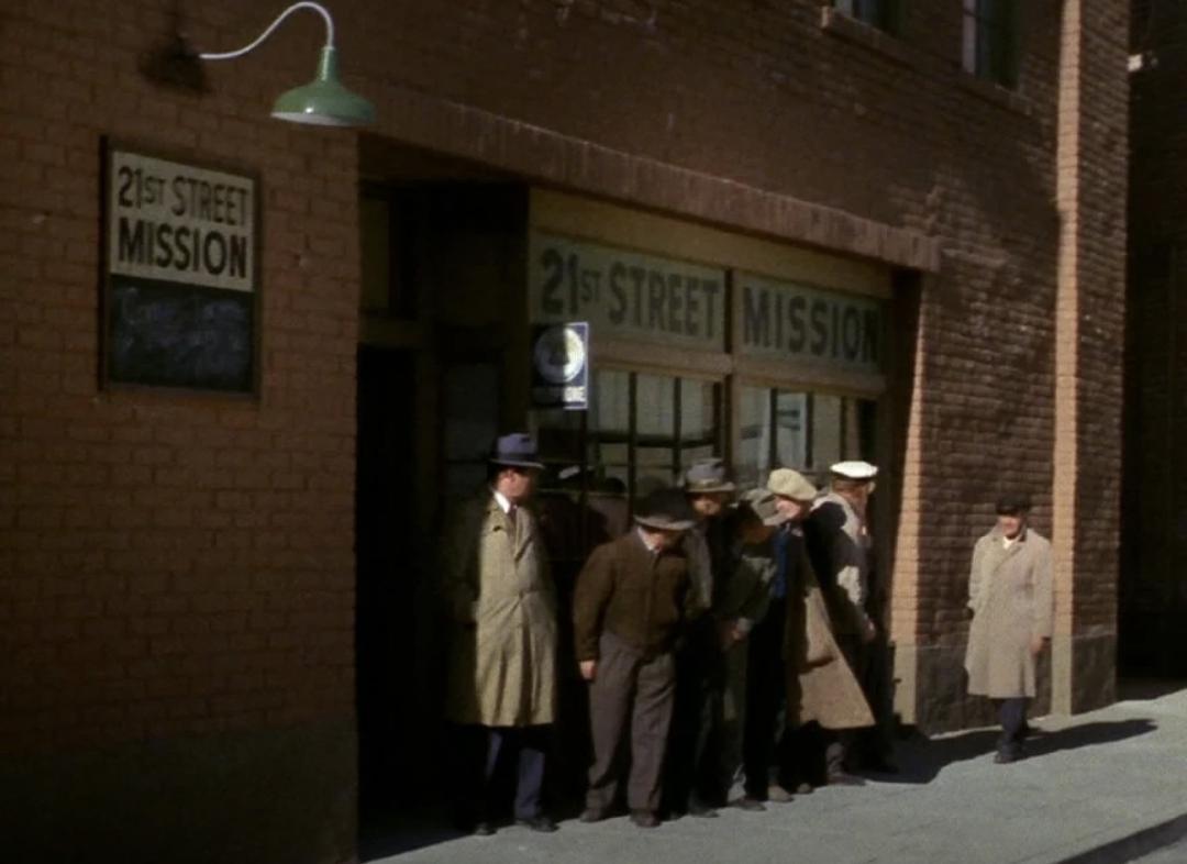 Outside (street view) of the 21st Street Mission in New York City. A number of men in hats and coats are lined up outside.