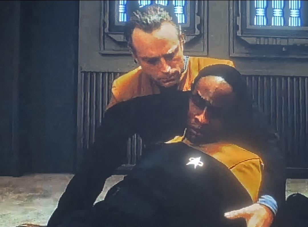 Suder cradling an unconscious Tuvok in his arms. He is looking at Tuvok with concern.