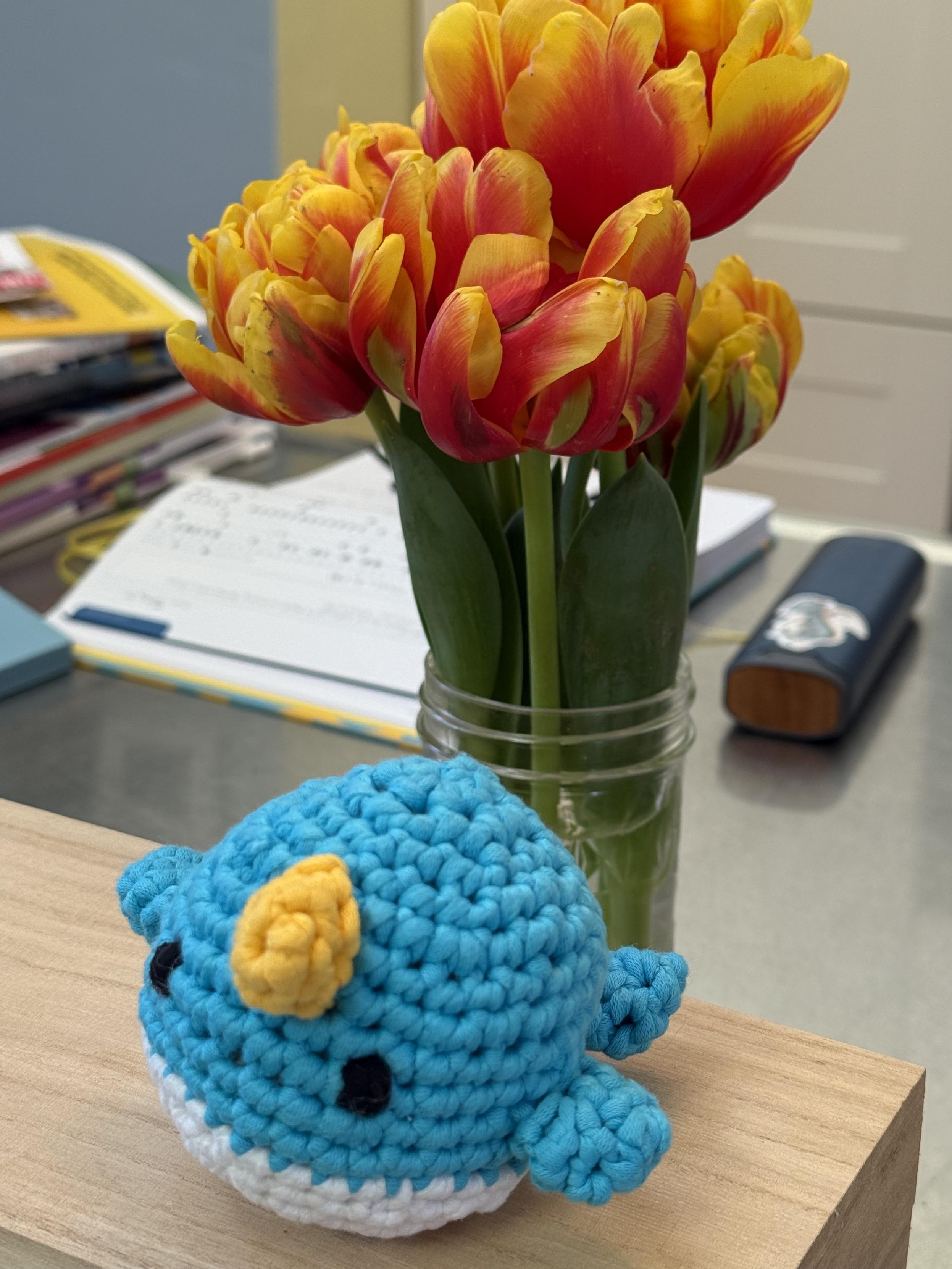 A lopsided crocheted narwhal in front of some tulips on a table.