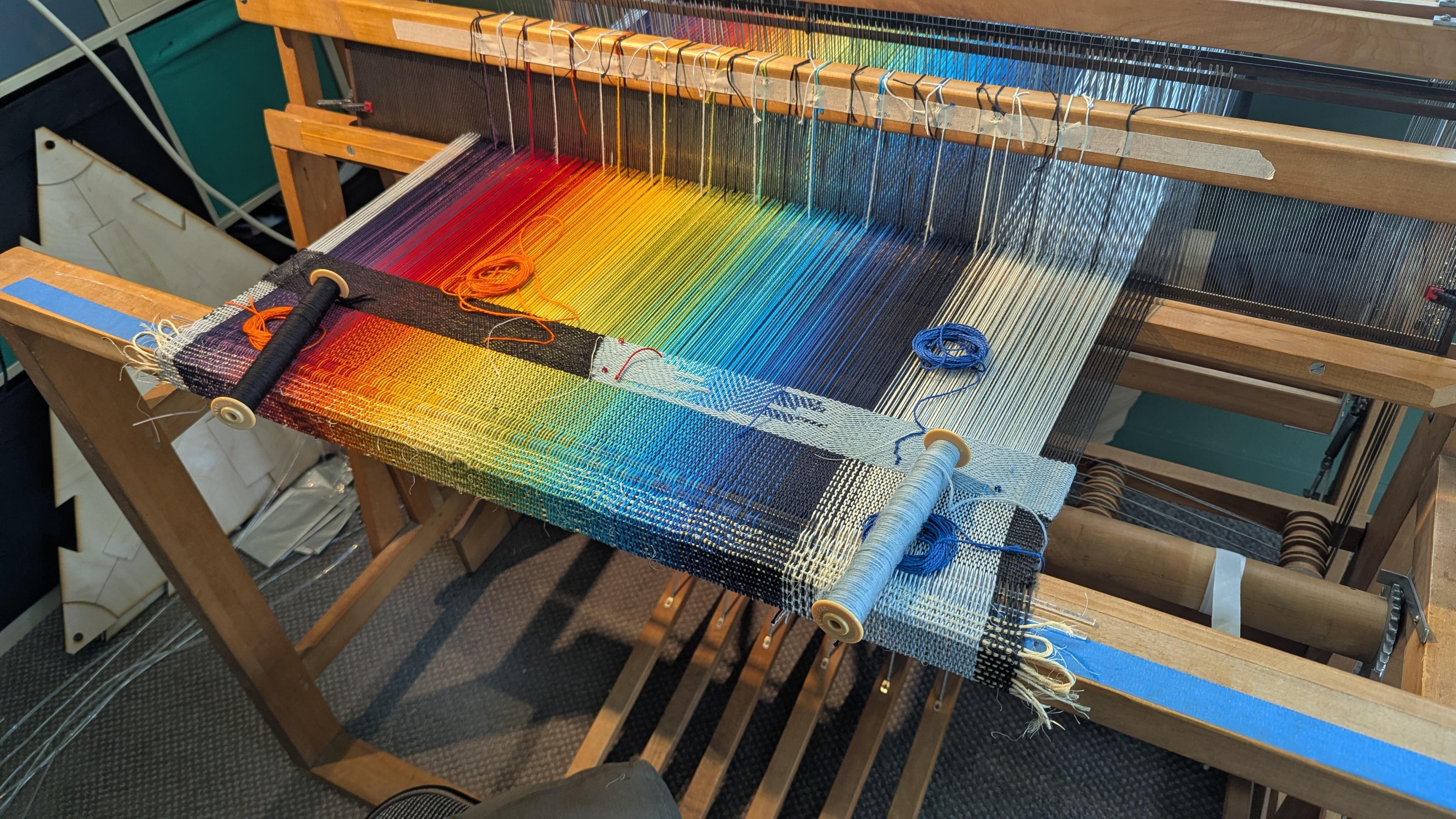 A floor loom set up to work, with a brilliant rainbow of colors in the warp: starting with dark purple on the left, into red and through basically the whole spectrum of colors to a navy blue near the right, where there's a sudden cutover to white and pale blue. A few inches of the material have already been woven, in black on the left and sky blue on the right, but there is a span in the mid-blue range where the warp color shows through. There are also a bunch of threads tied around the reed at regular intervals, and extra threads all over the surface, some on bobbins and some just floating loose.