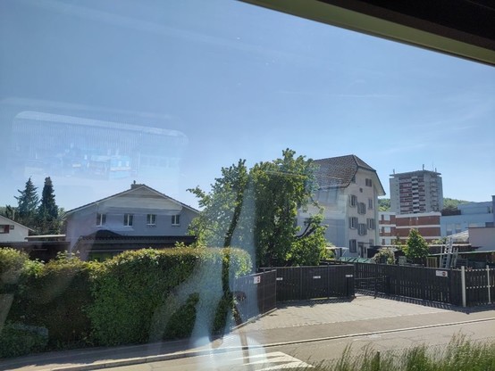 Foto aus einem Zugfenster, es sind eine Strasse und ein paar Häuser zu sehen. Eines davon hat ein Krüppelwalmdach.