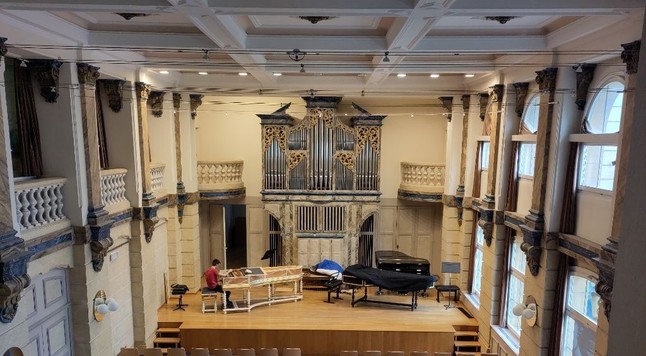 Foto eines hohen Saales mit einer weissen Kassettendecke, einer grossen Orgel und einer Bühne mit verschiedenen Musikinstrumenten