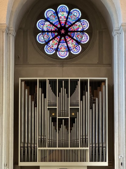 A large pipe organ with numerous metallic pipes is situated beneath an ornate, circular stained glass window featuring vibrant colors and intricate designs.