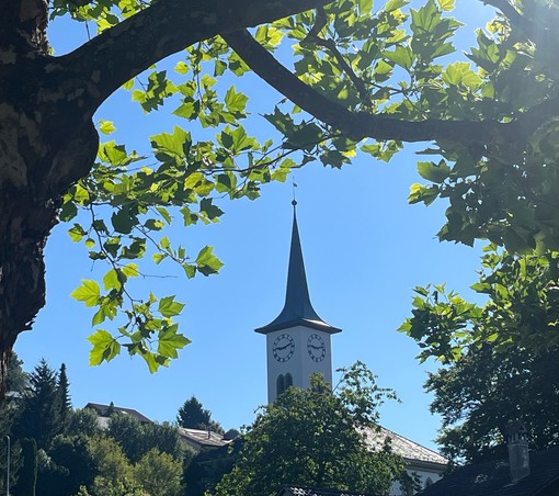 Umrahmt von grünen Blättern (Baum im Vordergrund) und vor blauem Himmel ein Kirchturm. Das Zifferblatt zeigt 9.10 Uhr