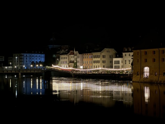 Der Landhausquai in Solothurn am späten Abend. Es ist dunkel. Der Landhausquai ist durch eine Lichterkette hell erleuchtet, deren Licht auf dem Wasserspiegel der Aare reflektiert.